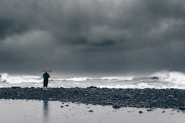 man on the shore near the sea, ocean in a raincoat and with an umbrella during a storm, hurricane,...