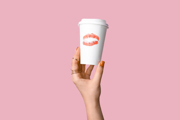 Female hand with red manicure holding paper cup of coffee on pink background