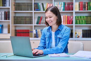 Young female university student studying inside library, typing on laptop computer