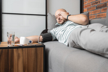 Unhealthy young man suffering from hypertension and headache, lying on the sofa at home, measures...