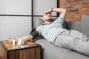 Unhealthy young man suffering from hypertension and headache, lying on the sofa at home, measures...