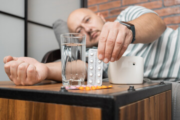 Obraz na płótnie Canvas Young adult man lying on a sofa at home measures blood pressure on a hand with modern digital tonometer and holding blister pack with pills