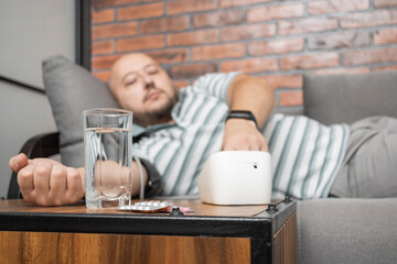 Defocused background with man who has hypertension and headache, lying on sofa at home, measures blood pressure on hand with modern digital tonometer, focus on blister pack with pills on foreground