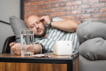 Obraz premium Defocused background with man who has hypertension and headache, lying on sofa at home, measures blood pressure on hand with modern digital tonometer, focus on blister pack with pills on foreground