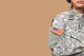 Confident young female soldier in uniform with American flag patch on sleeve saluting against beige...