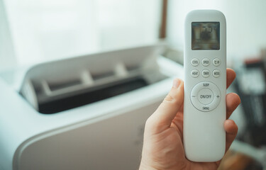 Hand holding remote control aimed at floor standing mobile air conditioner.