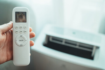 Hand holding remote control aimed at floor standing mobile air conditioner.