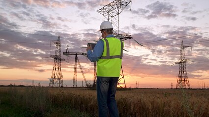Electrician in helmet checks data on tablet walking to power transmission lines