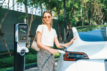 Young woman travel with EV electric car charging in green sustainable city outdoor garden in...