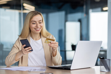 Young joyful woman at workplace inside office holds phone , bank credit debit card in hands, businesswoman chooses gifts in online store, book service and transfer funds using app on smartphone