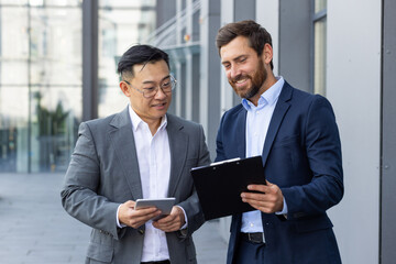 Two interracial business men standing outside an office building with a tablet and documents in...