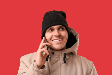Young man in stylish puffer jacket and hat talking by mobile phone on red background