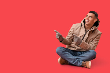Young man in stylish puffer jacket and gloves pointing at something on red background