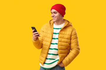 Young man in stylish puffer jacket and hat with mobile phone on yellow background