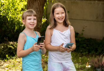 Happy school age kids two young girls children holding their smartphones mobile phones in hands outdoors, outside smiling. Using modern technology, apps, games, texting abstract concept, real people