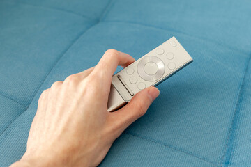 Man picking up holding a modern silver smart TV remote in his hand on a couch, sofa watching television, online video streaming smart TV equipment and usage, media consumption simple abstract concept