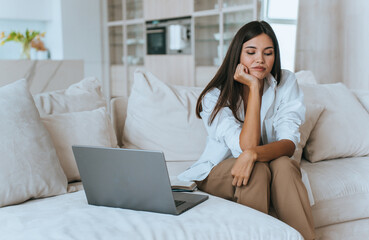 bored brunette is sitting at home on couch with a laptop looking down leaning on her hand tired and feeling apathy. Upset young girl feels lonely, upset, did not enter the institute
