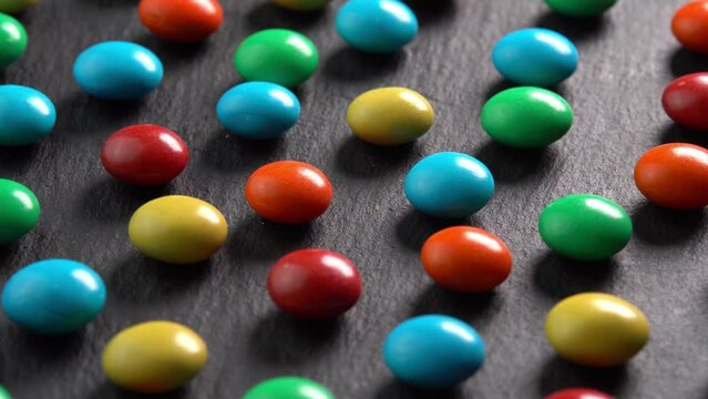 Sweet dragees in colored glaze rotating on black plate background macro side view sun light. Delicious sweets and candies close-up that children like. Chocolate Coating