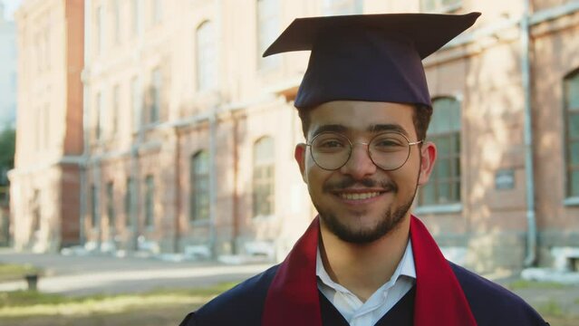 Medium close up portrait of cheerful holder of masters degree in hat and gown looking at camera outdoors