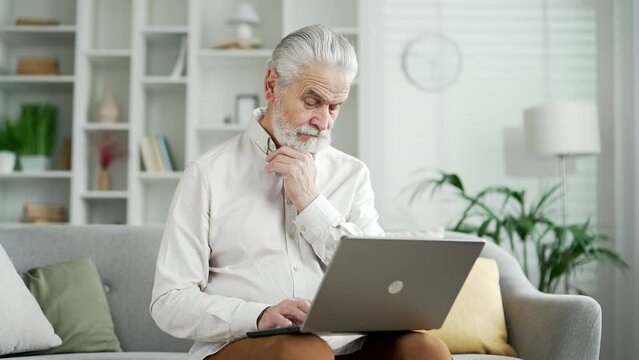 Serious thoughtfu elderly senior businessman working on laptop sitting on sofa in living room at home. Mature male texting messaging on computer browsing surfing web, online chatting banking in app