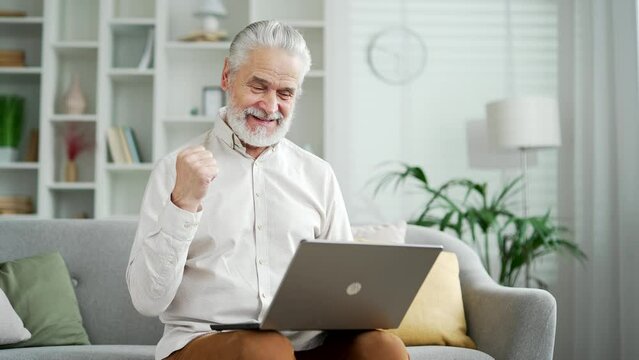 Happy excited elderly senior man with gray hair received great news on laptop sitting on sofa in living room at home. Smiling adult mature male reads a pleasant message on computer, celebrates success
