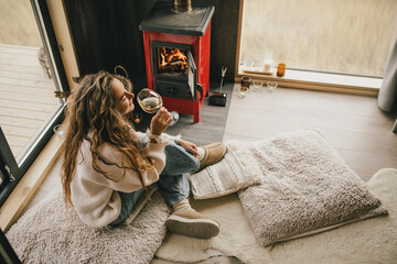 Young woman sitting by the fireplace in white sweater, drinking wine in cozy log cabin. - obrazy, fototapety, plakaty