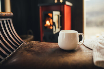 White cup with tea with burning fireplace on the background in cozy log cabin.