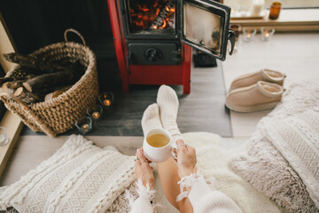 Fototapeta na wymiar Young woman sitting by the fireplace in white sweater, drinking tea in cozy log cabin.