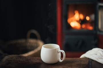 White cup with tea with burning fireplace on the background in cozy log cabin. - obrazy, fototapety, plakaty