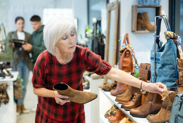 Senior woman shopper in leather product store finds it difficult to choose and thoughtfully view...