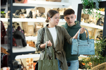 Young man with girl friend inspects shop window and is looking for modern, fashionable and roomy shoulder bag. In leather goods store, customers make choice of product with reasonable price - obrazy, fototapety, plakaty