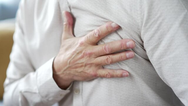 Close up. Mature male hands holds massaging chest. Sick elderly senior man suffering from chest tension, heart attack sitting on the sofa in the living room at home. He needs urgent medical attention