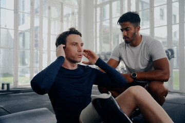 Multiracial trainer next to young man in sportswear with artificial limb doing push-ups at the gym
