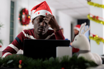 Depressed exhausted african american employee struggling with demanding company project during Christmas holiday season. Unhappy stressed worker getting overwhelmed by tasks in festive ornate office - obrazy, fototapety, plakaty