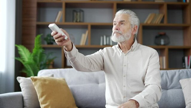Elderly senior man with gray hair turns on air conditioner by remote control sitting on sofa in living room at home. Mature male presses the button of the device and turns off the heating in the house