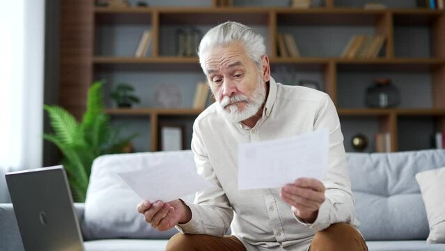 Frustrated elderly senior gray haired man looking at large utility bills sitting on sofa at home. Upset mature male holding paycheck, thinking about problems in household budget and financial expenses