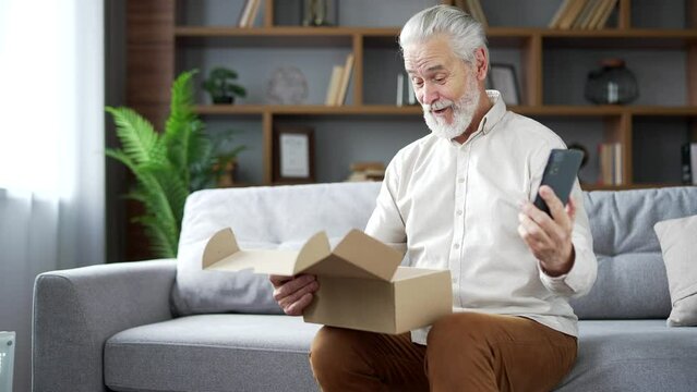 Elderly senior man receiving a parcel with a gift at home talking online via video call using smartphone. Smiling joyful male is happy with shopping in an online store. Satisfied client of e-commerce
