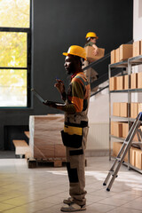 African american employee analyzing inventory checklist on laptop computer, looking at carton boxes full with customers orders in warehouse. Supervisor working at merchandise delivery in storage room