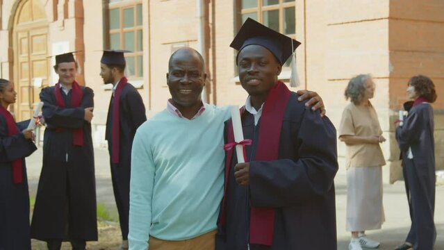 Portrait of young African American man in gown and hat posing for photo together with his father against university