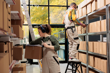 Supervisor carrying cardboard box analyzing checklist while working in storage room, preparing clients orders. Storehouse employee wearing headphones listening music during inventory