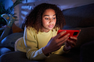 Young latina girl looking concentrated at mobile lying on sofa in living room. Serious afro woman watching cell phone video at night home. Female generation z black people and domestic leisure time