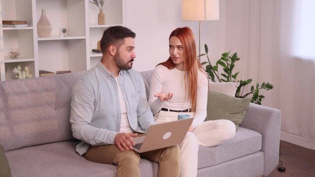 Happy smiling young family couple talking sitting on the couch with laptop computer browsing product in online internet store planning apartment decoration and imagine their beautiful house together