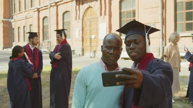 Medium shot with selective focus on male bachelor taking selfie with his father on smartphone during graduation day