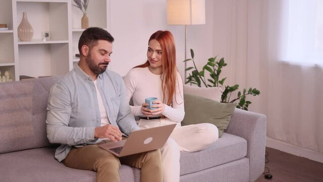 Happy smiling young family couple talking sitting on the couch with laptop computer browsing product in online internet store planning apartment decoration and imagine their beautiful house together