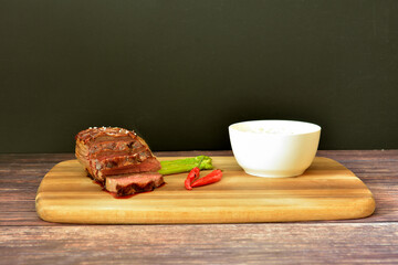 Grilled beef steak with asparagus and hot peppers on a wooden rack, next to a cup with sauce and cutlery.