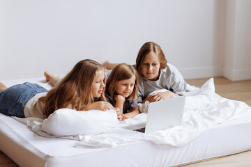 Three beautiful teenage girls are lying on the bed with a laptop and surfing the Internet. Sisters or girlfriends spend time together.