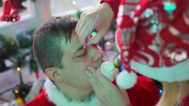 Girl doing make-up on a guy's face preparing for Christmas, a young couple dressed in festive costumes of Santa Claus and the Snow Maiden against the backdrop of Christmas decorations, close up.
