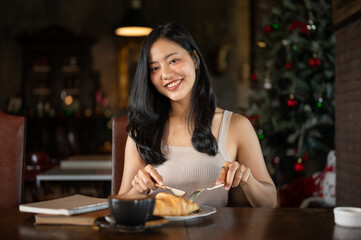 Elegant young Asian woman eating croissant fro breakfast with hot latte coffee in a dark luxury living room.