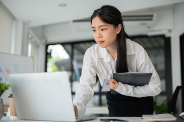 Young beautiful Asian businesswoman working for her client in her office room with laptop and office supplies.