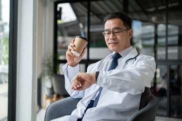 A senior male doctor is checking time on his wristwatch while taking a coffee break in his office.